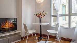 Una sala moderna con televisor de pantalla plana, una mesa redonda con un florero, dos sillas blancas y un ventanal con cortinas.