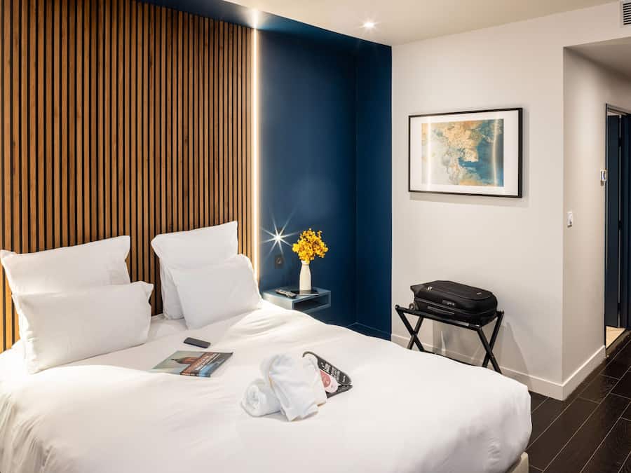 A hotel room with a neatly made bed, a bedside table with a vase of flowers, a framed map on the wall, and a suitcase on a trolley.