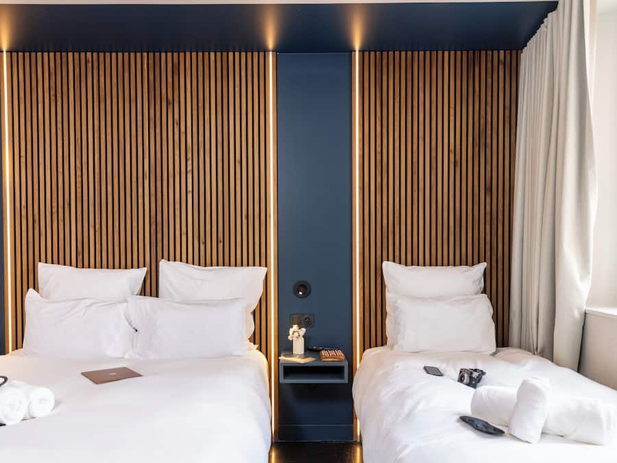 Ein modernes Hotelzimmer mit zwei Betten, Holzvertäfelung an den Wänden, einem Nachttisch mit Lampe und einem Fenster mit Vorhängen.