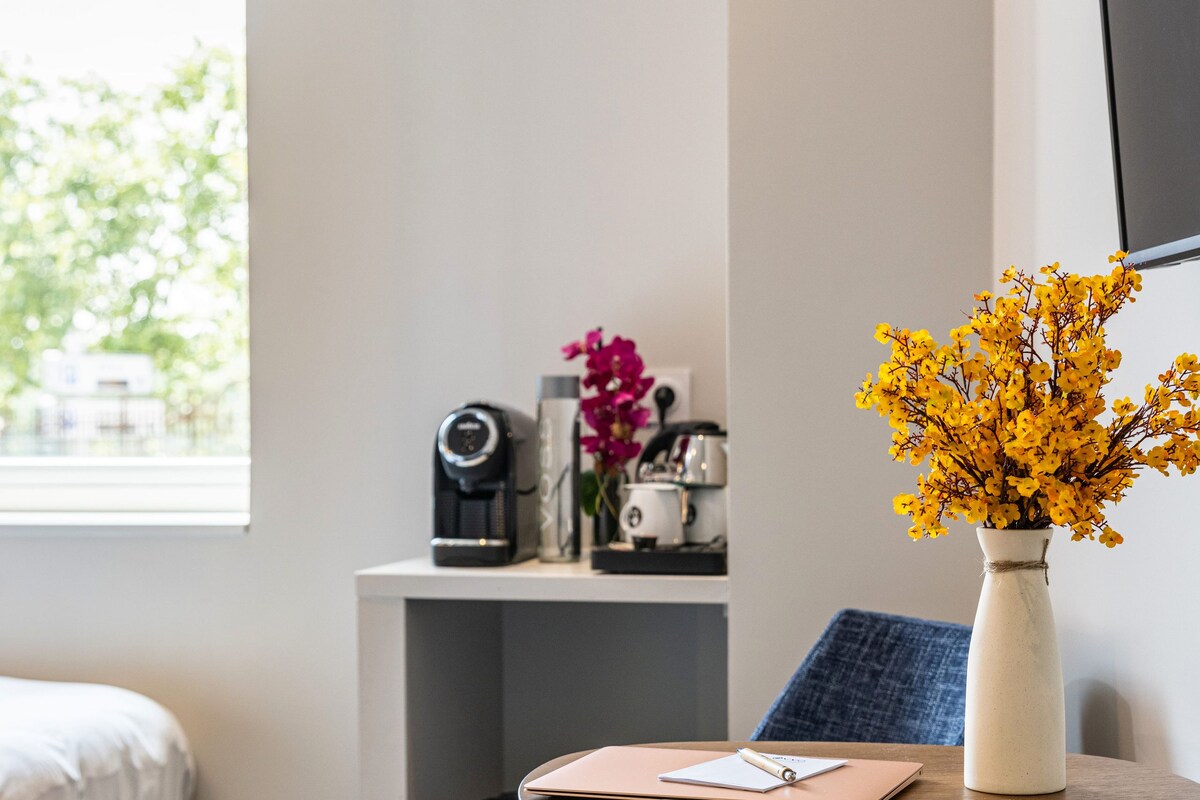 A small room with a bed, a desk with a vase of yellow flowers, a chair, and a table with a notebook and pen.