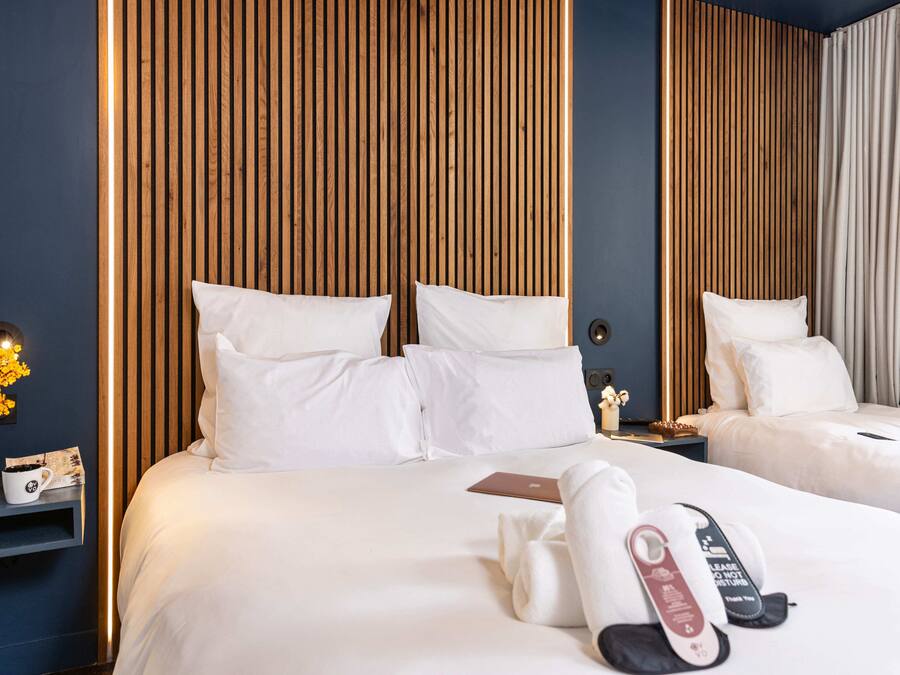 A hotel room with two beds, wooden slat headboards, a bedside table with a vase of flowers, a remote control, and a pair of slippers.