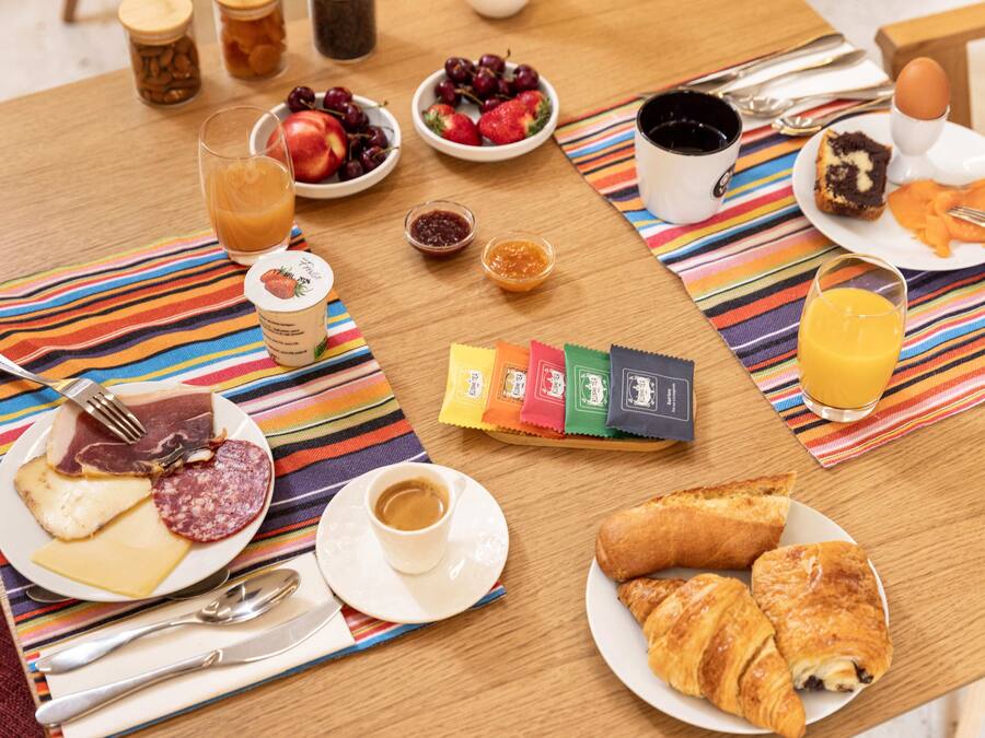 Una mesa de desayuno con platos de comida, incluyendo cruasanes, queso y fruta, además de un vaso de zumo de naranja y una taza de café.