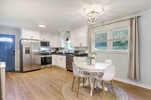 Una cocina moderna con armarios blancos, electrodomésticos de acero inoxidable, una mesa redonda con sillas y una ventana con cortinas.