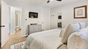 Un dormitorio con una cama, ventilador de techo, televisión, cómoda y un baño visible a través de una puerta abierta.