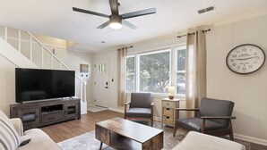 Una sala de estar con un televisor de pantalla plana, una mesa de centro, dos sillones, un sofá, un ventilador de techo y un reloj grande en la pared.