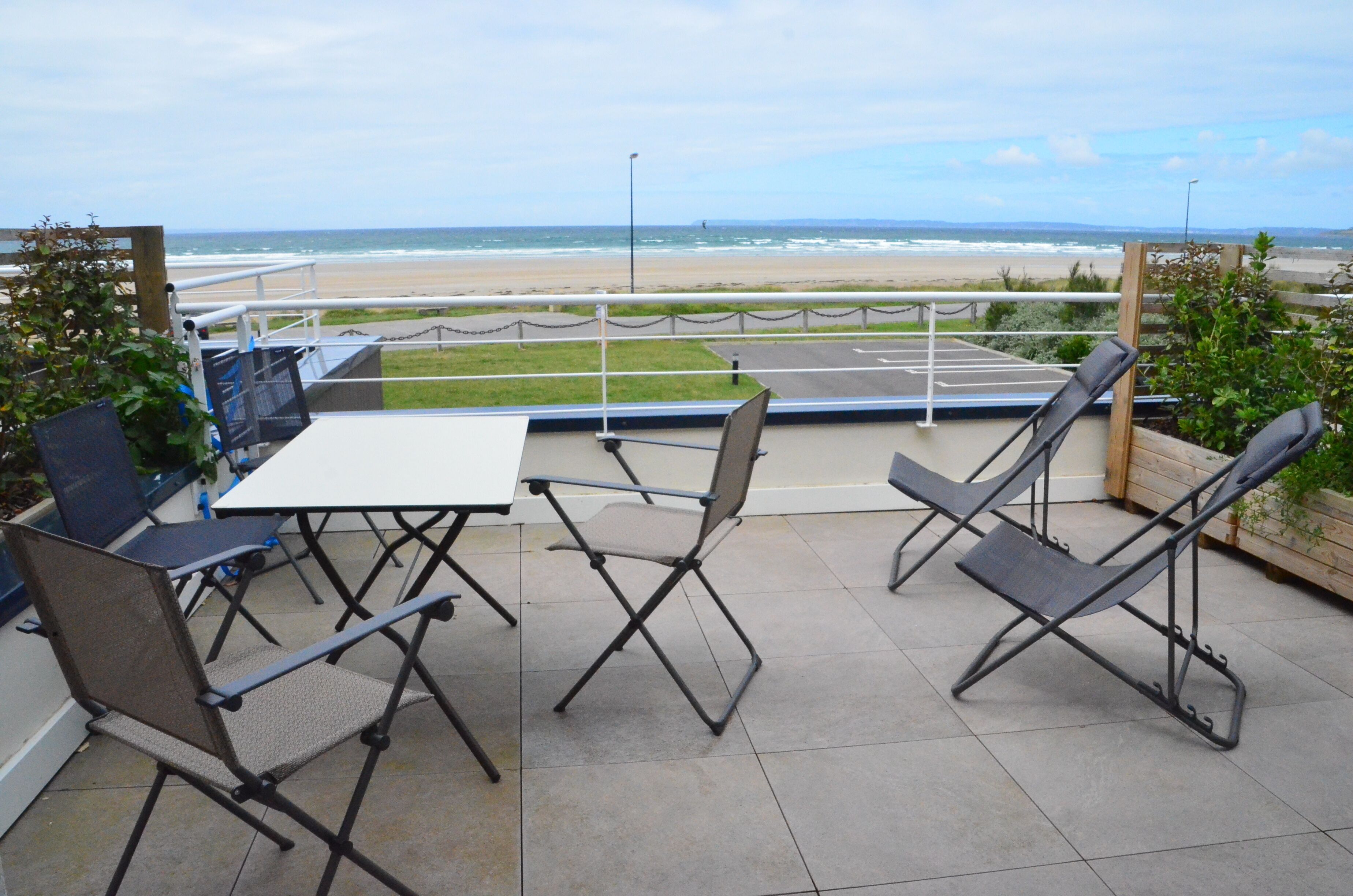 Une terrasse extérieure avec une table et des chaises, une vue sur la plage et un espace de stationnement.
