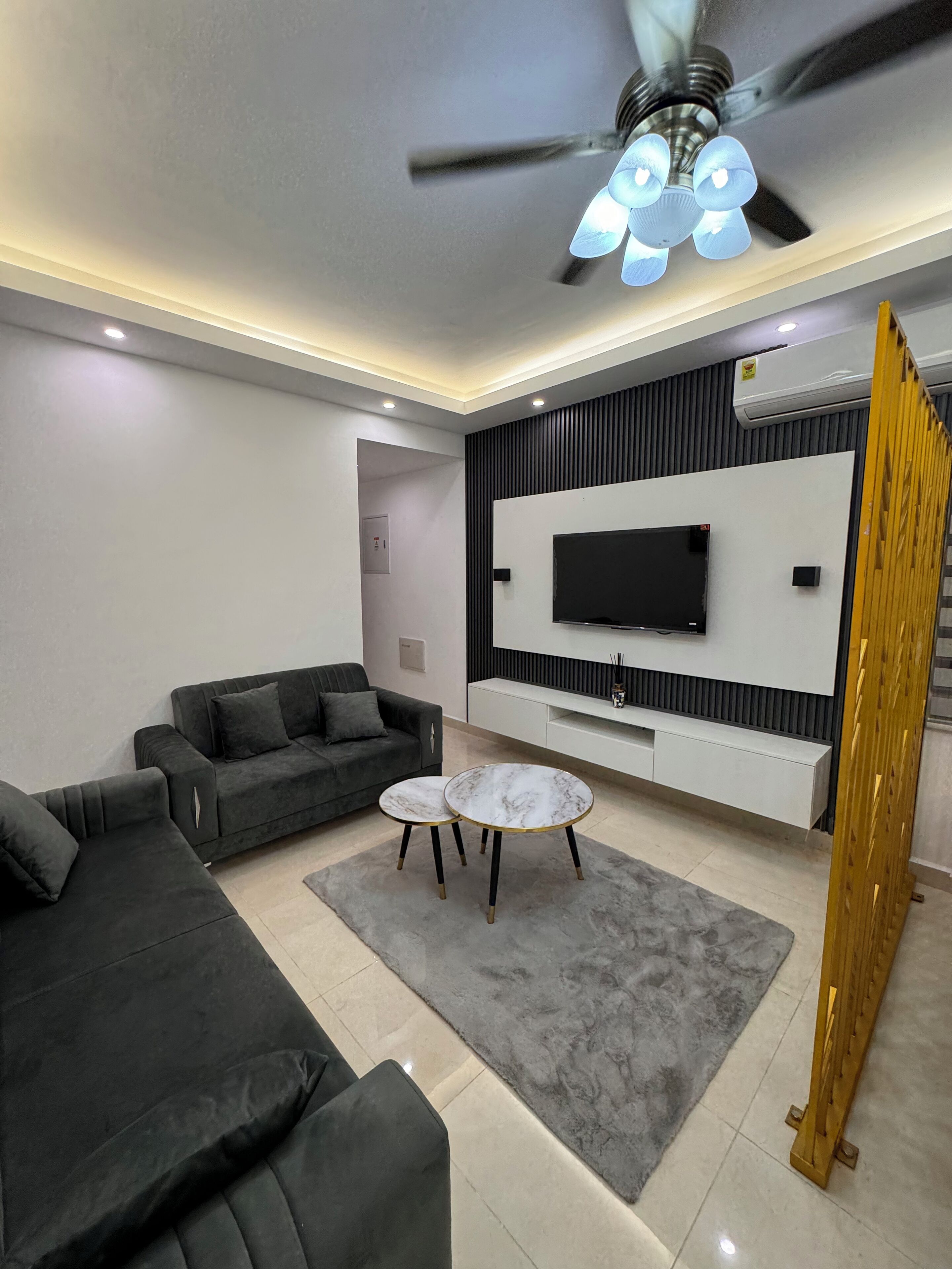 Una sala de estar moderna con un conjunto de sofá, una mesa de centro, un televisor montado en la pared y un ventilador de techo.