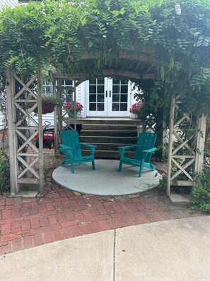 A wooden pergola with two green chairs under a white door, surrounded by greenery.