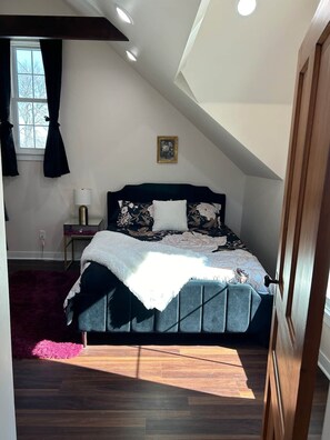 A bedroom with a sloped ceiling, a bed with a floral bedspread, a bedside table with a lamp, and a window with curtains.