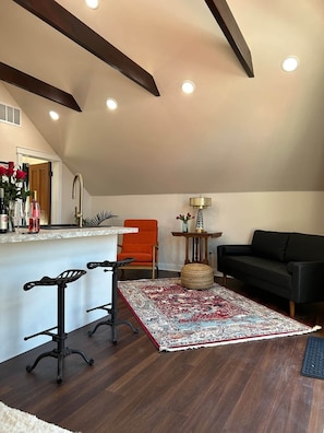 A modern living room with a black leather sofa, a red chair, a dining table with chairs, a patterned rug, and a kitchen area with a sink and countertop.