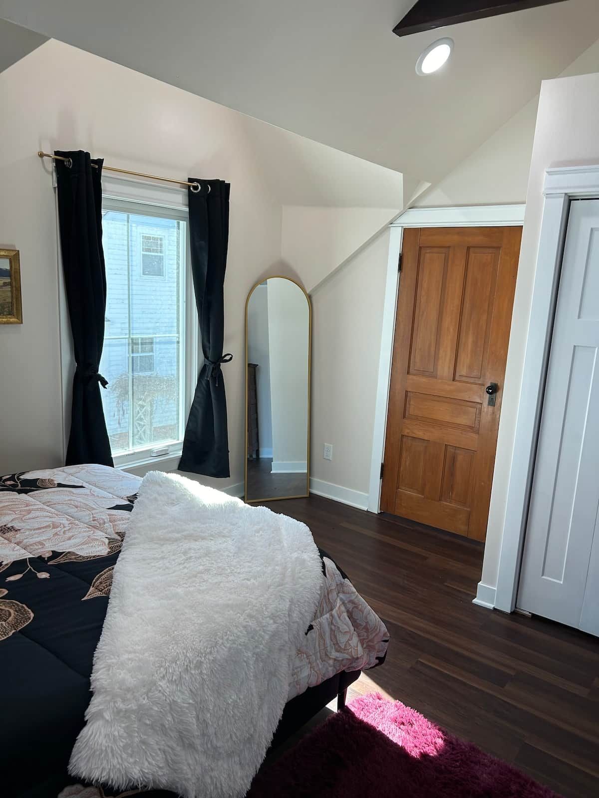 Un dormitorio con cama, puerta de madera, espejo y ventana con cortinas.