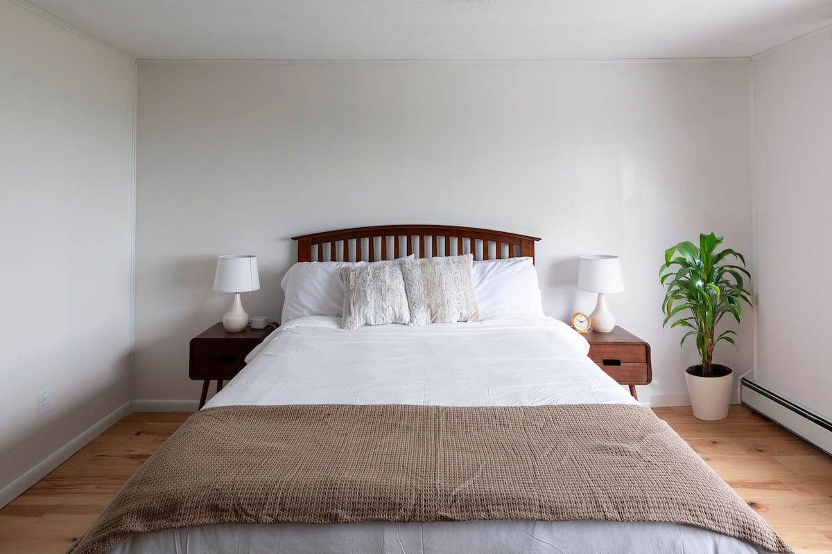 A neatly made bed with a wooden headboard, two bedside tables with lamps, a potted plant, and a brown throw blanket.