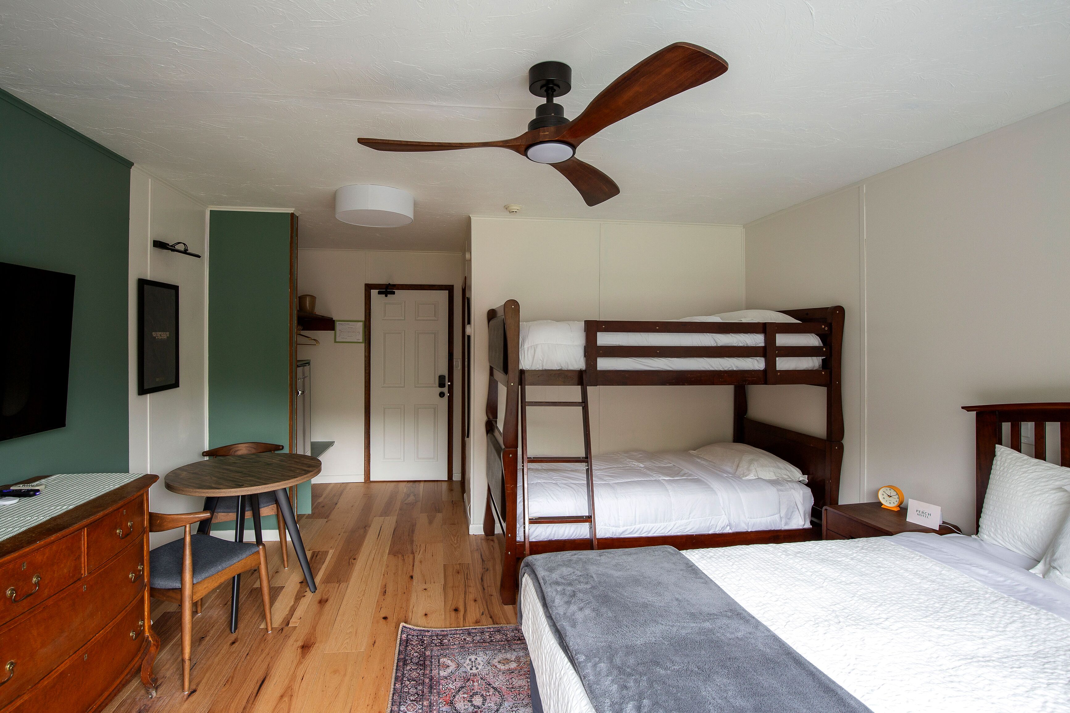 A room with bunk beds, a desk with a chair, a television, and a ceiling fan.