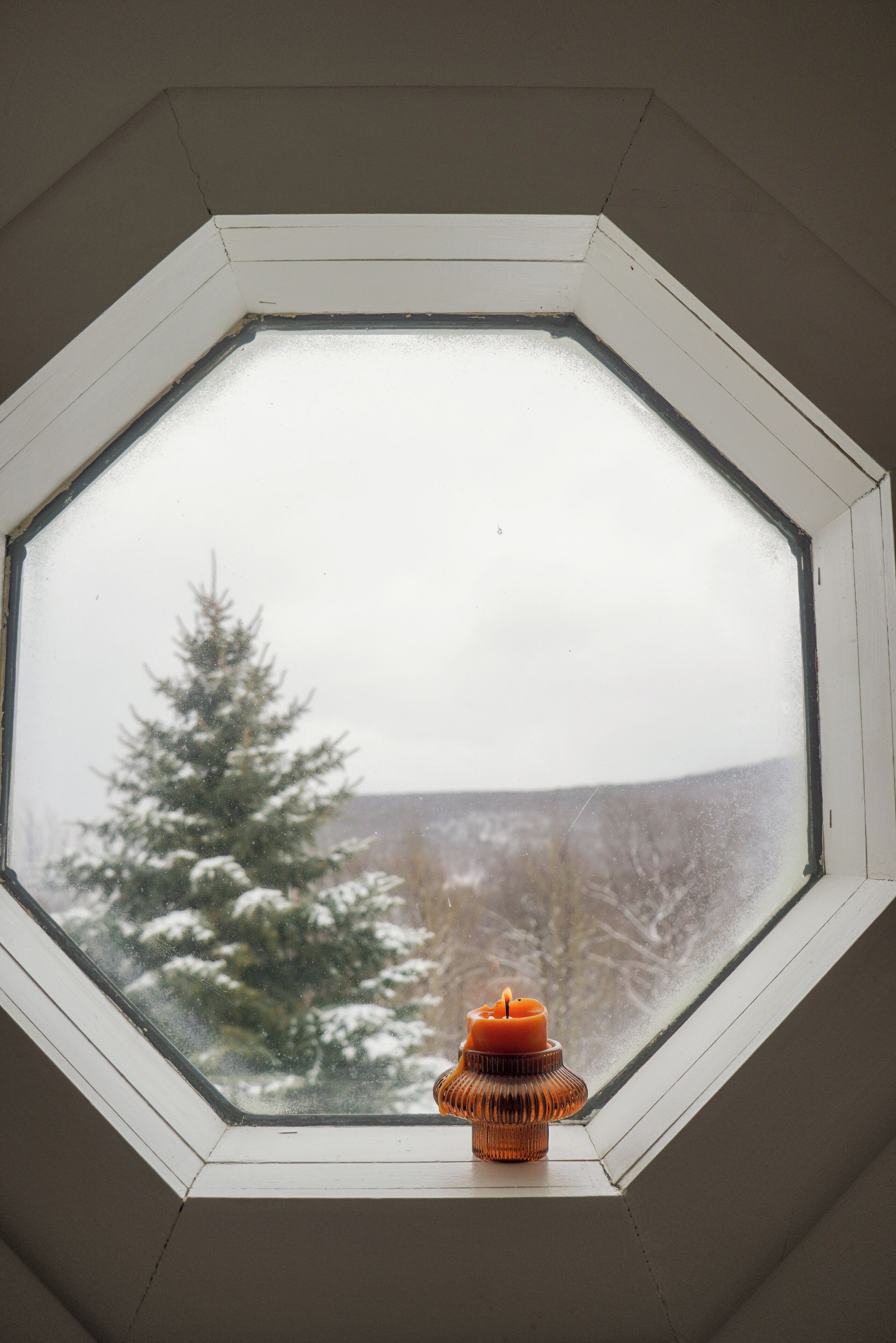 Un tragaluz con una vela en su interior, que da a un árbol cubierto de nieve y un paisaje lejano.