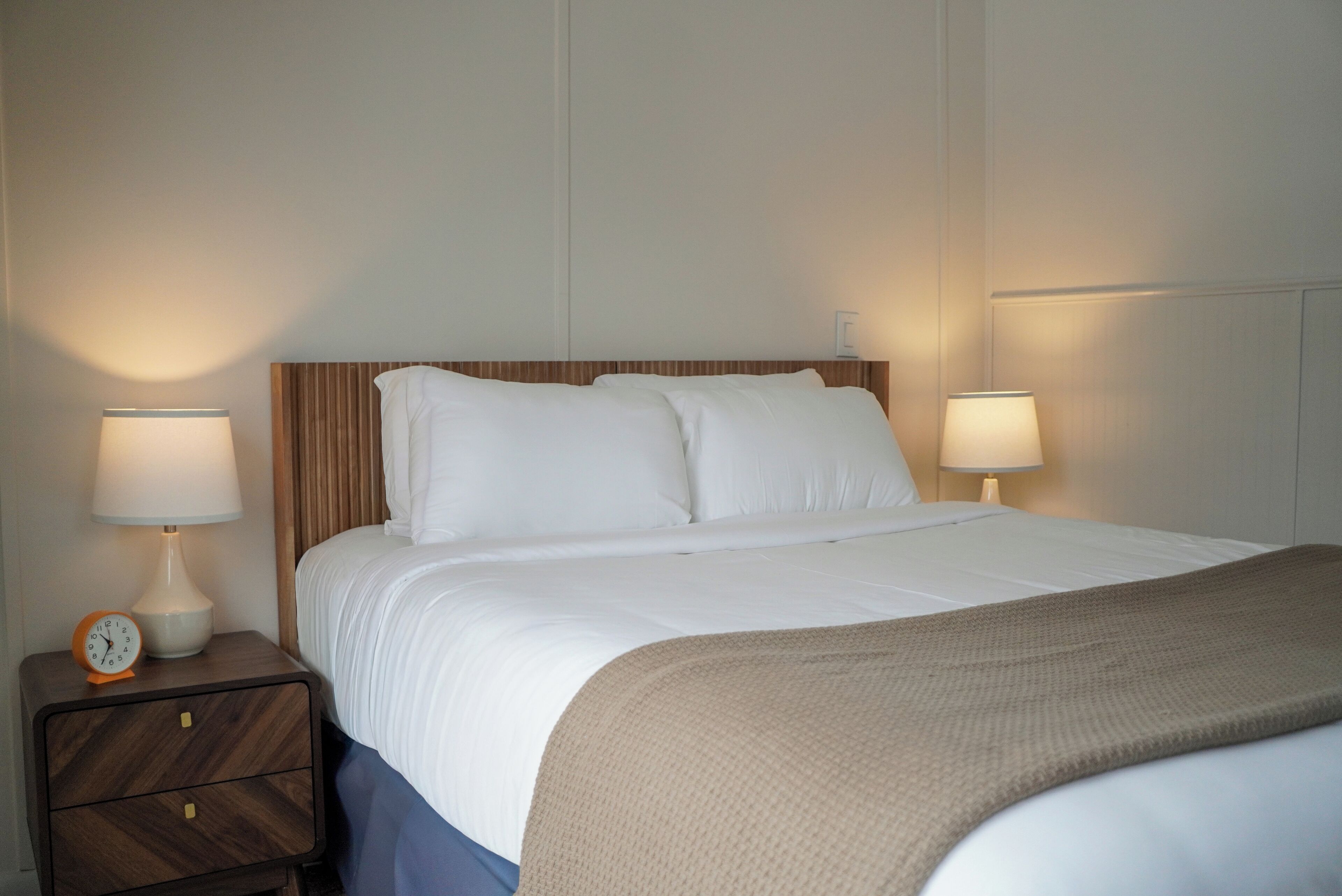 Een netjes opgemaakt bed met witte lakens en een houten hoofdbord, met aan weerszijden nachtkastjes met lampen en een klok.