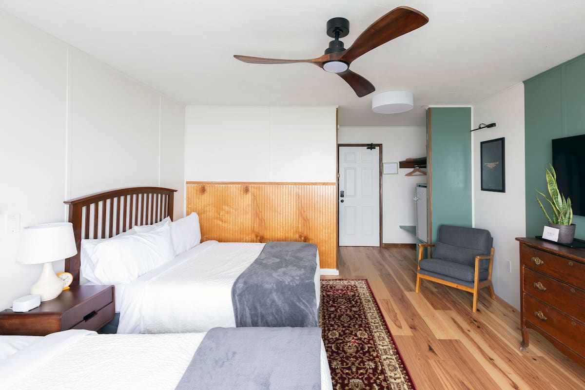 A hotel room with two beds, a wooden headboard, a ceiling fan, a desk with a lamp, a chair, a dresser, and a potted plant.