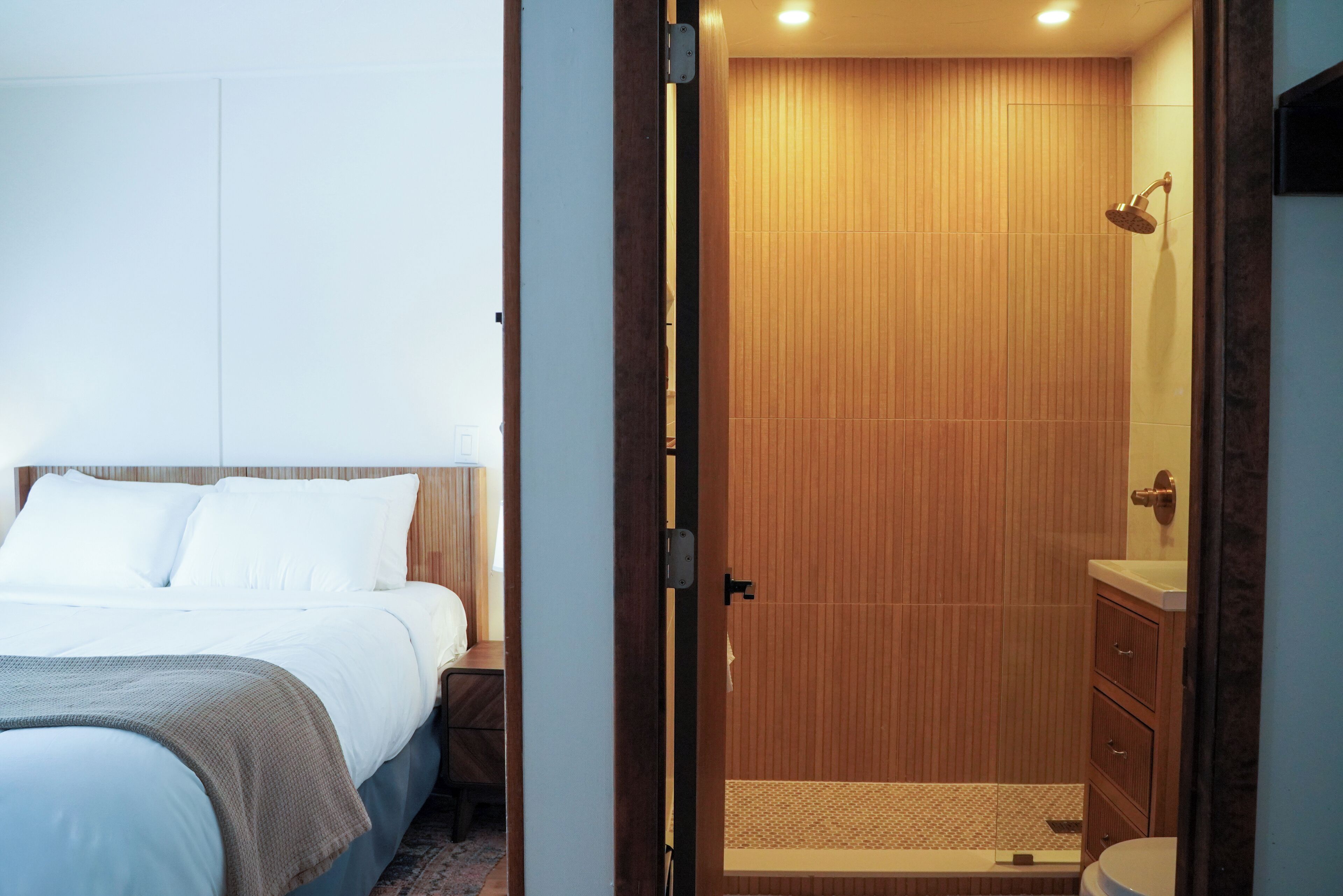 Quarto de hotel com uma cama, box de chuveiro em madeira e banheiro com pia e espelho.