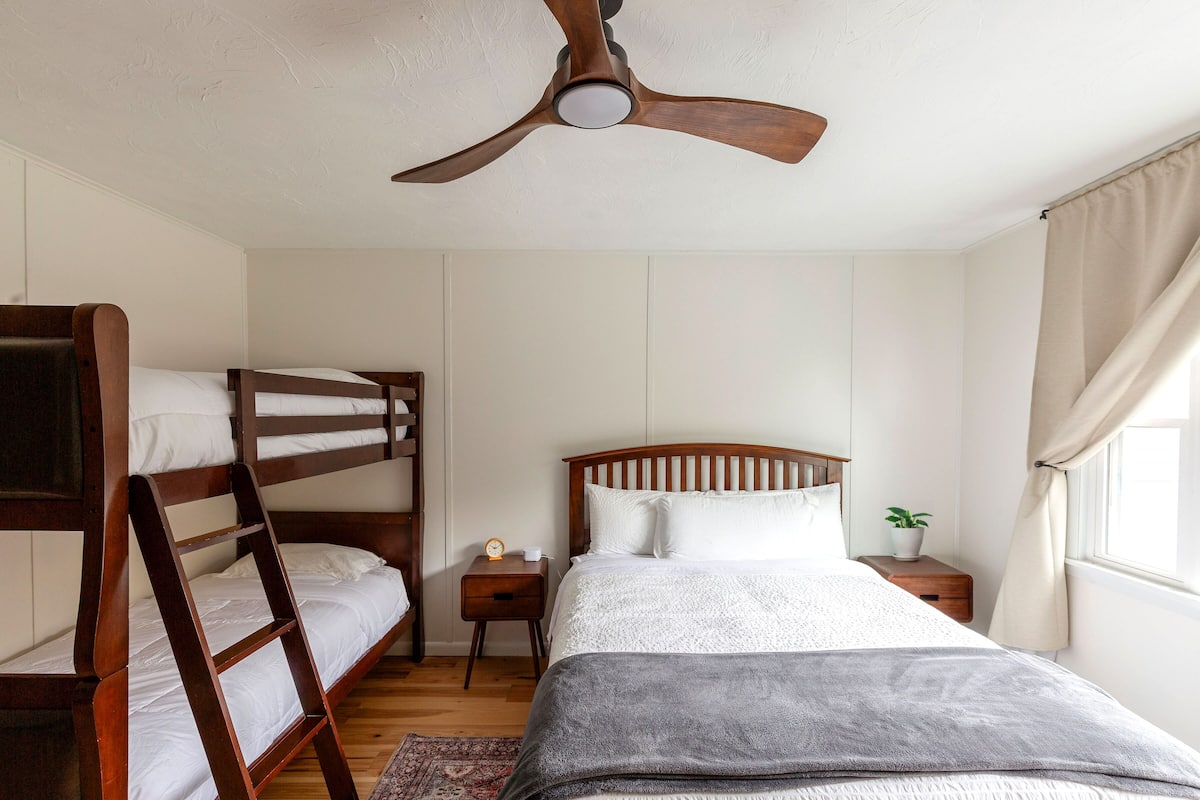 A bedroom with bunk beds, a single bed, a ceiling fan, a bedside table with a plant, and a window with curtains.