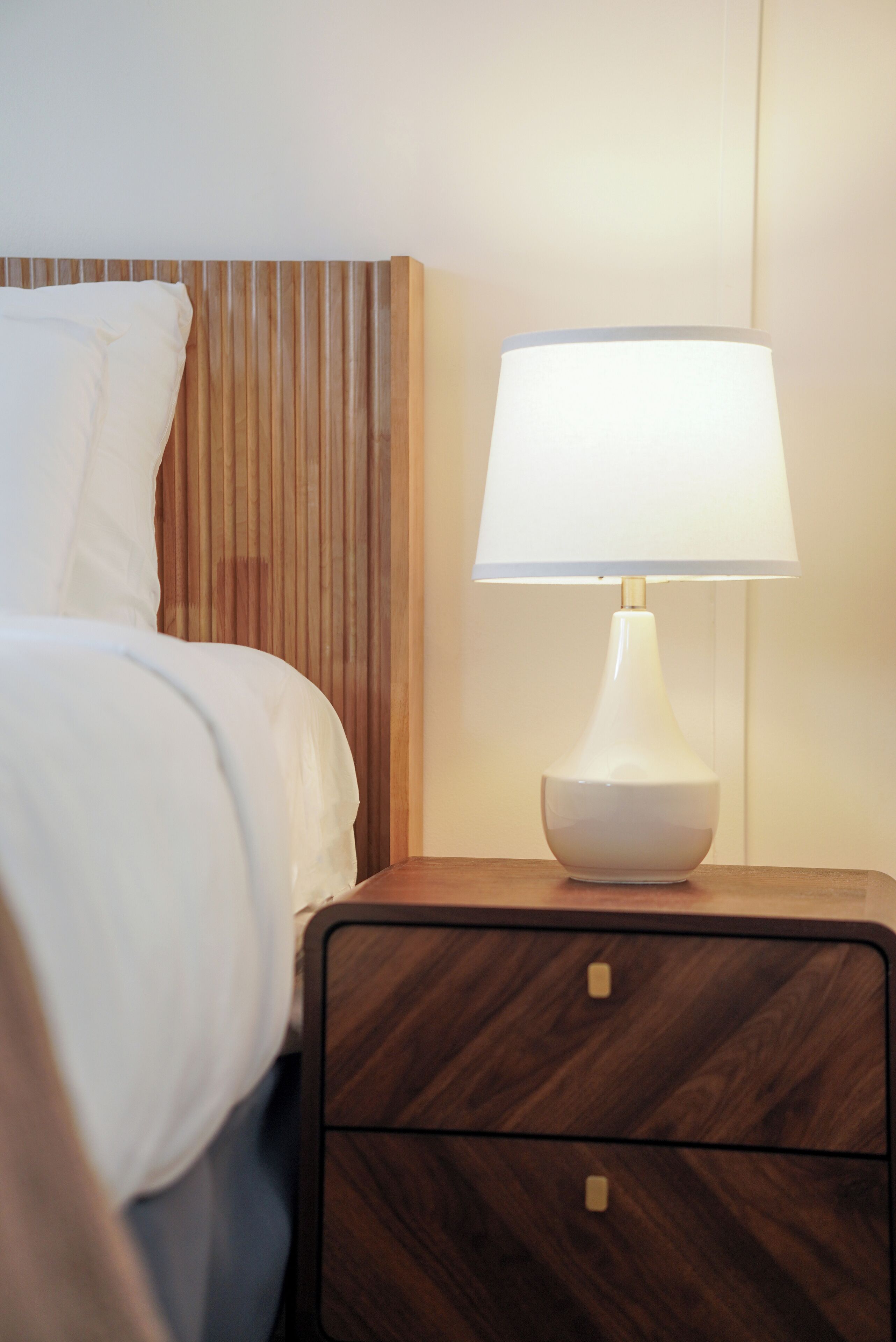 A neatly made bed with white linens, a wooden headboard, and a bedside table with a white lamp.