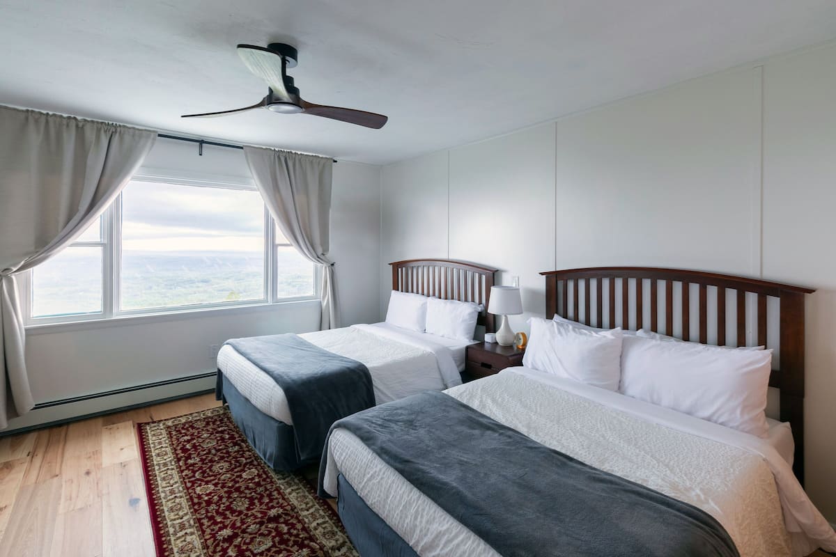 A hotel room with two beds, a ceiling fan, a nightstand with a lamp, and a view of the outdoors through the window.