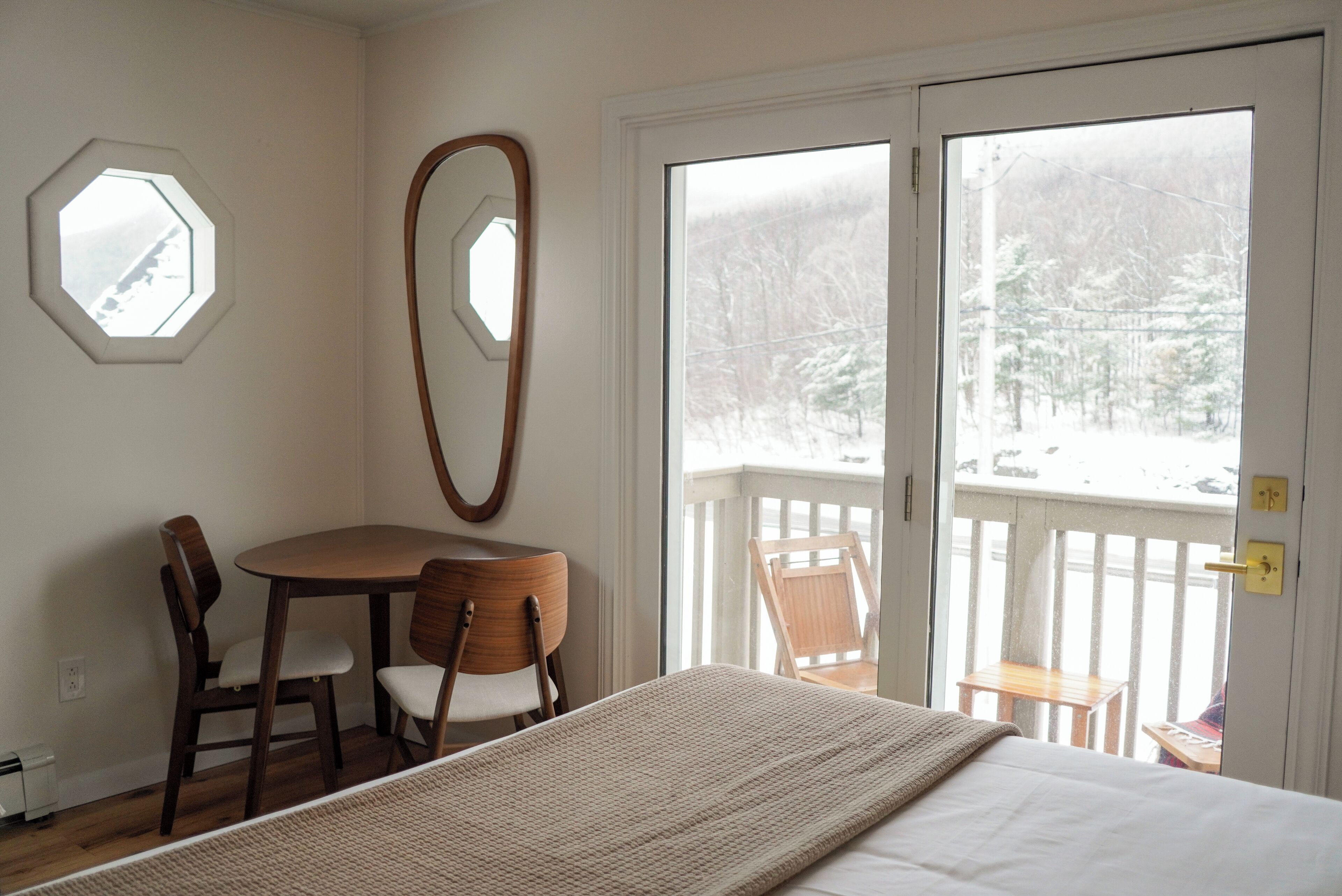 Una habitación con una cama, una mesa de madera con sillas, un espejo redondo y vistas a una terraza nevada a través de una puerta corrediza de vidrio.