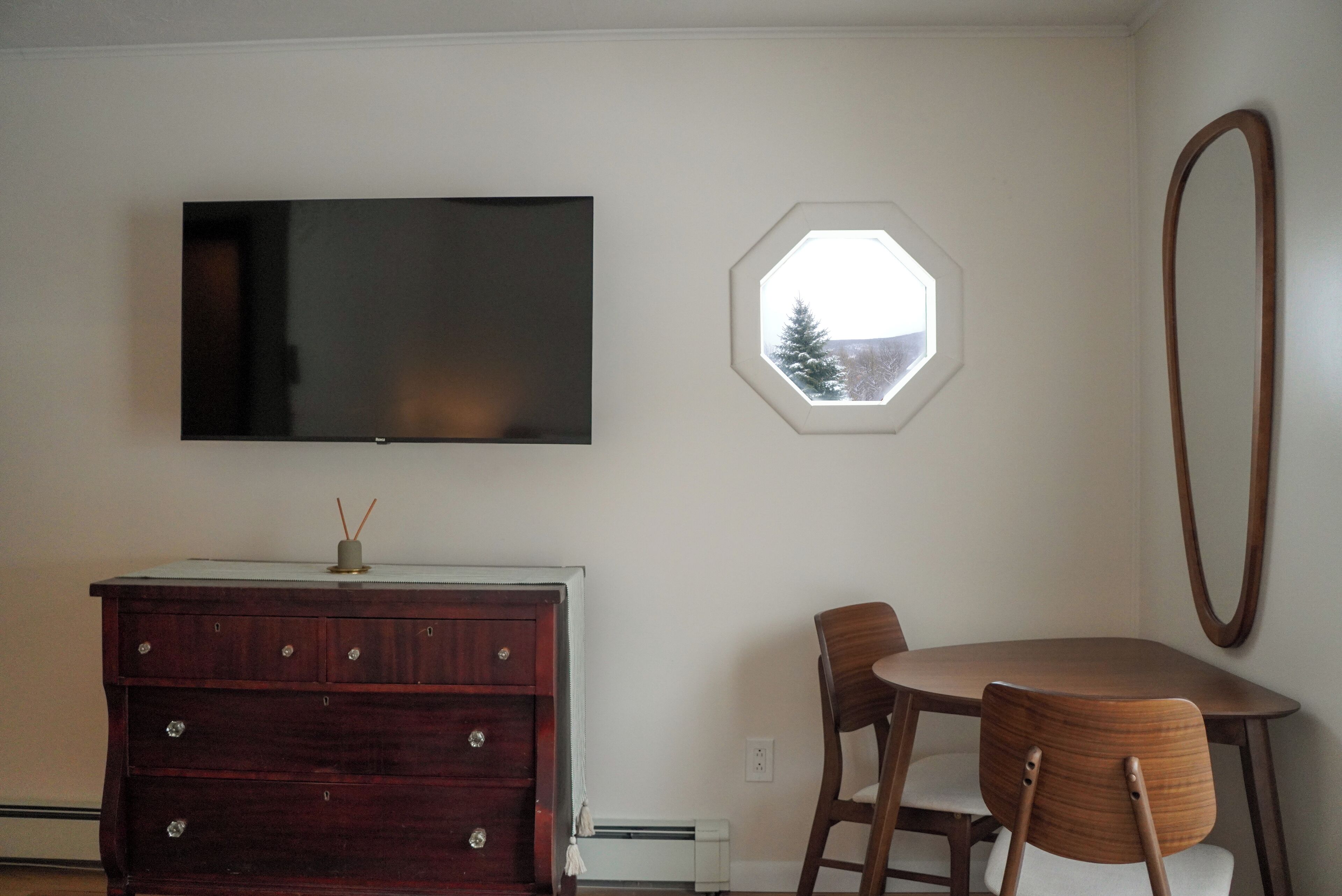Una habitación con una televisión de pantalla plana montada en la pared, un aparador de madera, una mesa redonda con sillas y una ventana con vista a árboles.