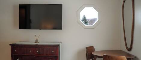 A room with a flat-screen TV mounted on the wall, a wooden dresser, a round dining table with chairs, and a window with a view of trees.