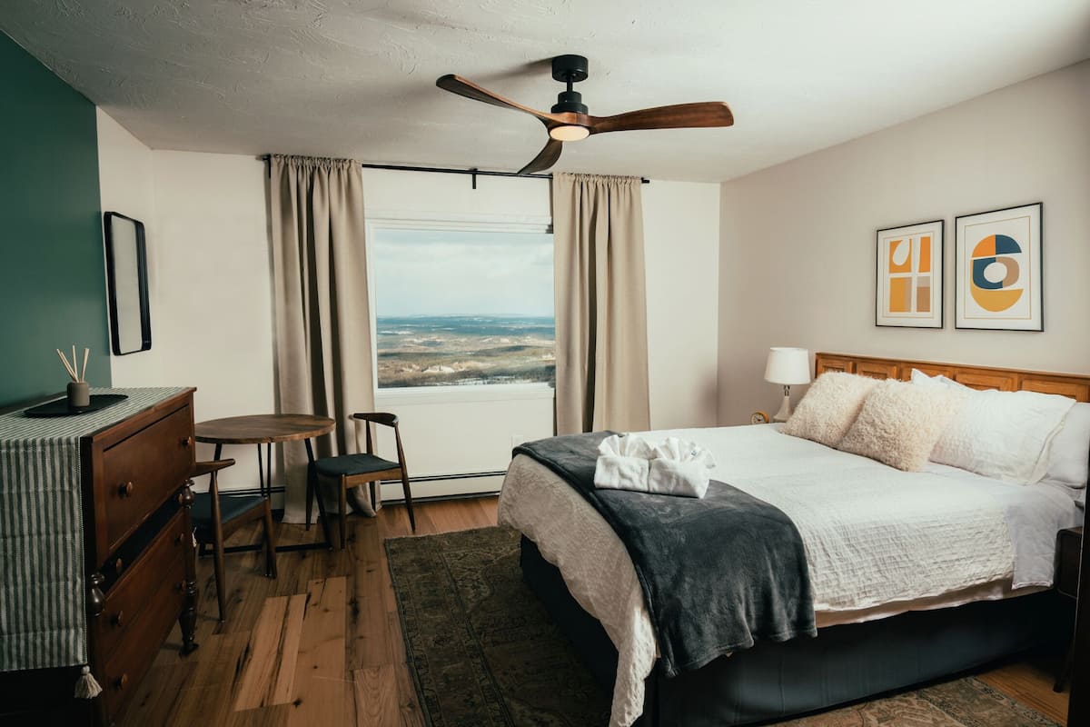 A hotel room with a bed, a desk, a chair, a small table, a ceiling fan, a window with curtains, and two framed pictures on the wall.