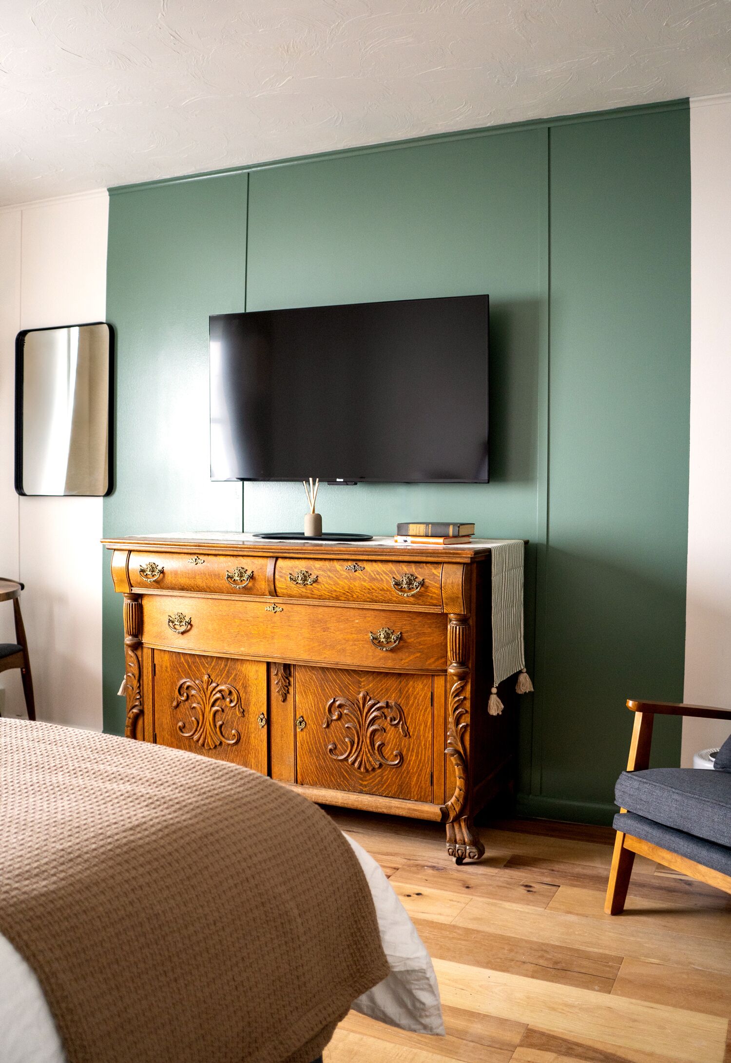 Een kamer met een houten dressoir, een aan de muur bevestigde televisie, een bed met een beige deken en een blauwe stoel.