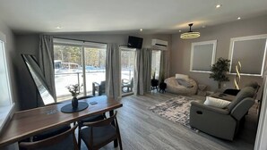 A modern living room with a sofa, a dining table with chairs, and a large window with curtains. - Treehouse Farm Stay on the Maine Coast (Harpswell)