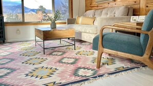 Ein gemütliches Wohnzimmer mit großem Fenster, Holzwänden, einem gemusterten Teppich und einer Couch mit Kissen.