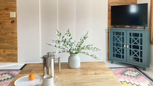 Ein Zimmer mit Holzwand, einem Fernseher auf einem Schrank, einem Schrank, einem Tisch mit Kaffeekanne, einer Vase mit Blumen und einem Teller mit einer Orange.