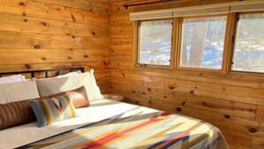 Ein Schlafzimmer in einer Holzhütte mit einem Bett, Kissen und einem Fenster mit Blick auf eine verschneite Landschaft.