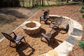 A stone fire pit with four chairs arranged around it in a backyard setting.