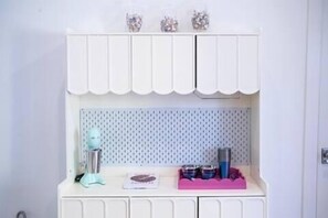 A white cabinet with a scalloped top, a perforated wall behind it, and a pink tray with items on the lower shelf.