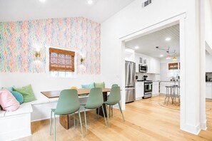 A modern kitchen with stainless steel appliances, a dining area with a wooden table and green chairs, and a colorful mosaic wall.
