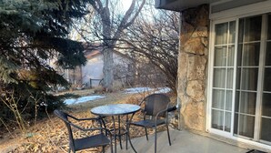 Un patio con dos sillas y una pequeña mesa, un muro de piedra, y vistas a una casa y árboles.