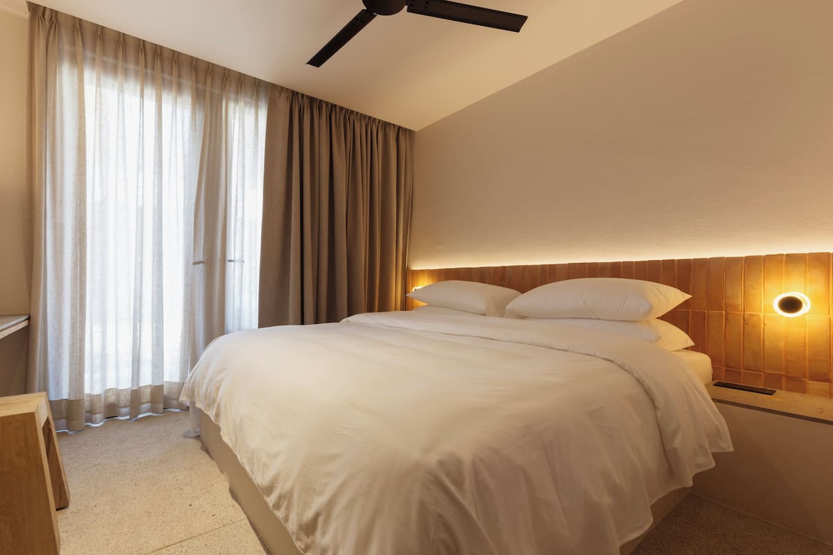 A neatly made bed with white linens, a wooden headboard, and a wall-mounted light fixture.