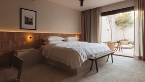 A bedroom with a bed, bedside table, bench, and a view of an outdoor area with plants.