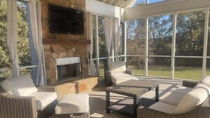 Un patio soleado con chimenea de piedra, asientos cómodos y ventanas amplias que ofrecen vistas al exterior.