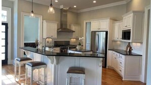 Una cocina amplia con armarios blancos, una isla central con taburetes, electrodomésticos de acero inoxidable e iluminación colgante.