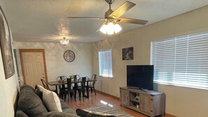 Ein Wohnzimmer mit einem Ecksofa, einem Esstisch mit Stühlen, einem Deckenventilator, einem Fernseher und einer Uhr an der Wand.