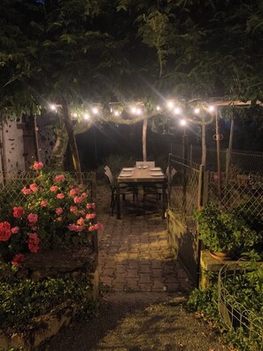 Ein Essbereich im Freien mit Tisch und Stühlen unter Lichterketten, umgeben von Grünpflanzen und einem Zaun.