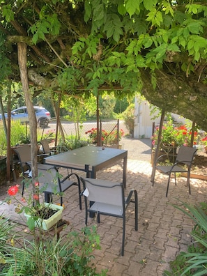 An outdoor seating area with tables and chairs under a tree, surrounded by greenery and flowers.