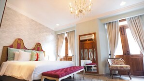 Ein Hotelzimmer mit einem großen Bett, einer Bank, einem Sessel, einer Kandelaber und einer Holztür.