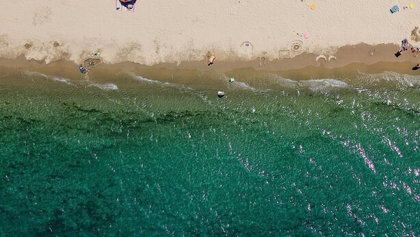 An aerial view of a beach with clear turquoise water, a sandy shore, and various beach umbrellas and chairs.