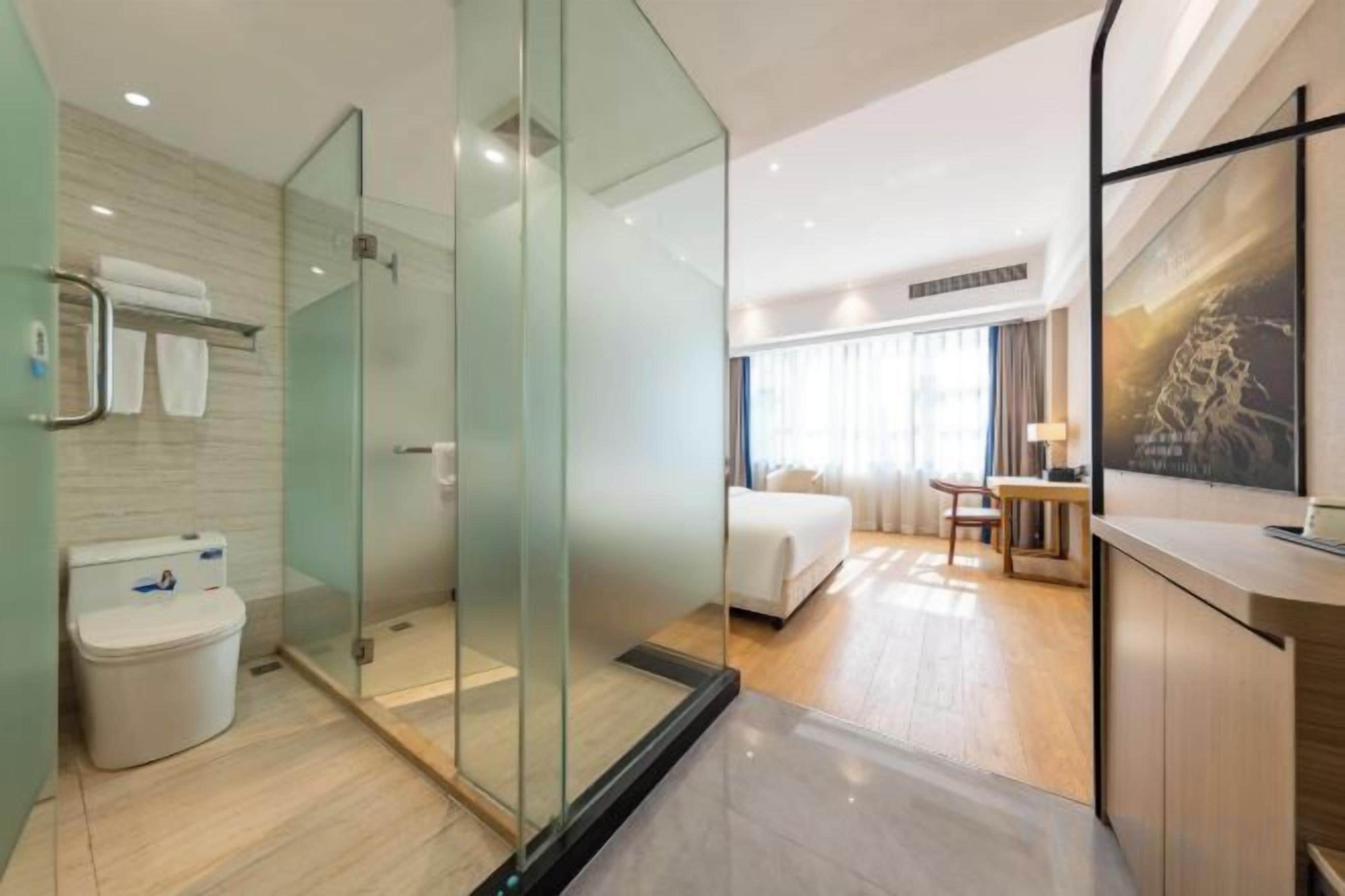 A modern hotel room with a glass-enclosed shower, a separate bathroom, a bed, a desk, and a large window.