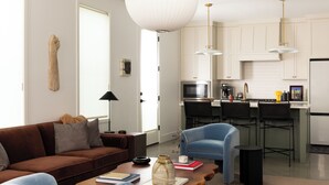 Un soggiorno moderno con un divano marrone, poltrone blu e un tavolino da caffè in legno. Si intravede in secondo piano una cucina con pensili bianchi e un forno a microonde.