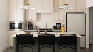 Una cucina moderna con mobili bianchi, un frigorifero in acciaio inossidabile, un forno a microonde e una macchina da caffè. Sono presenti tre sgabelli neri e una grande isola con una ciotola sopra.