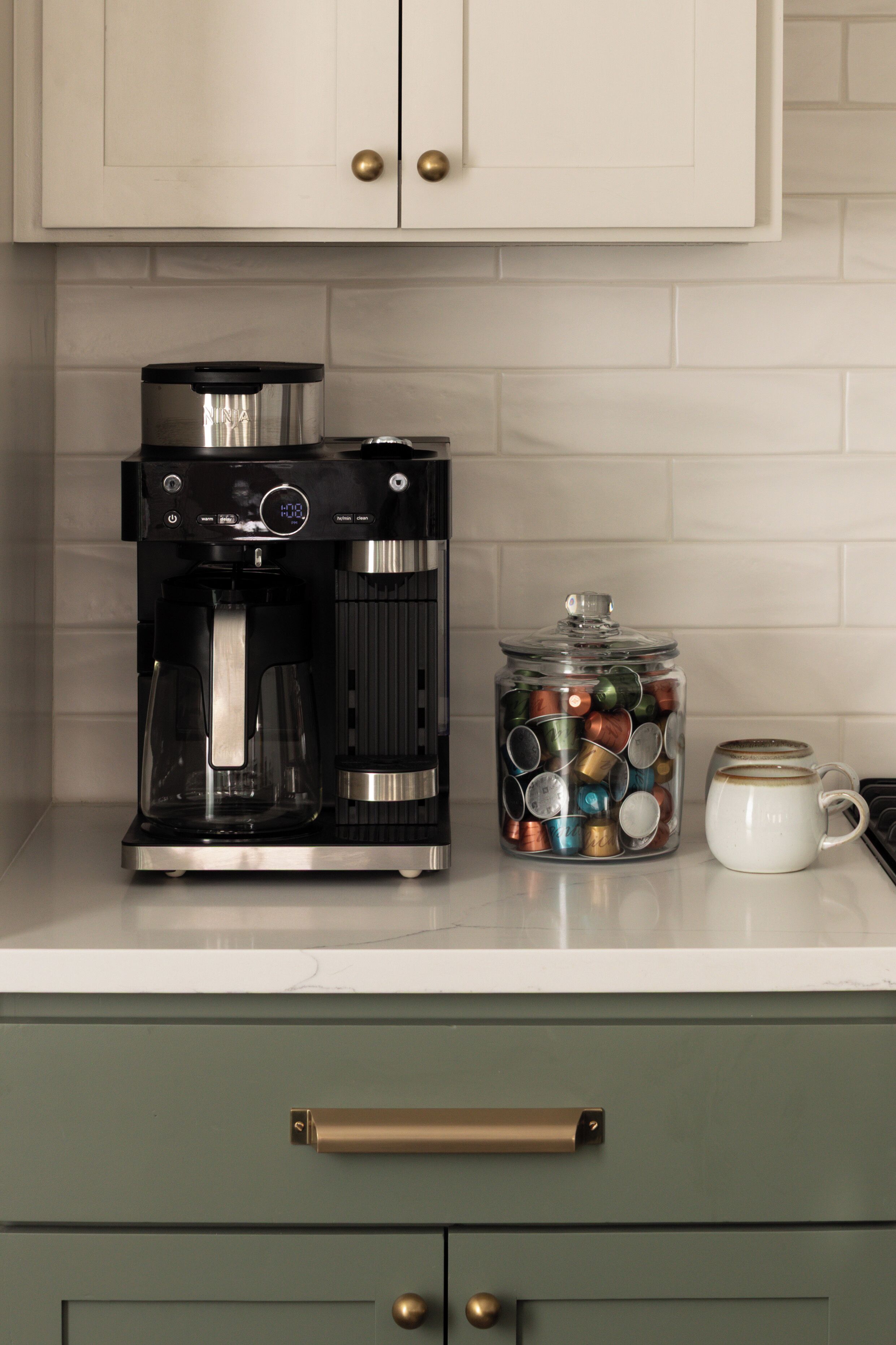 Una cafetera, un tarro lleno de cápsulas de café y una taza sobre una encimera de cocina.